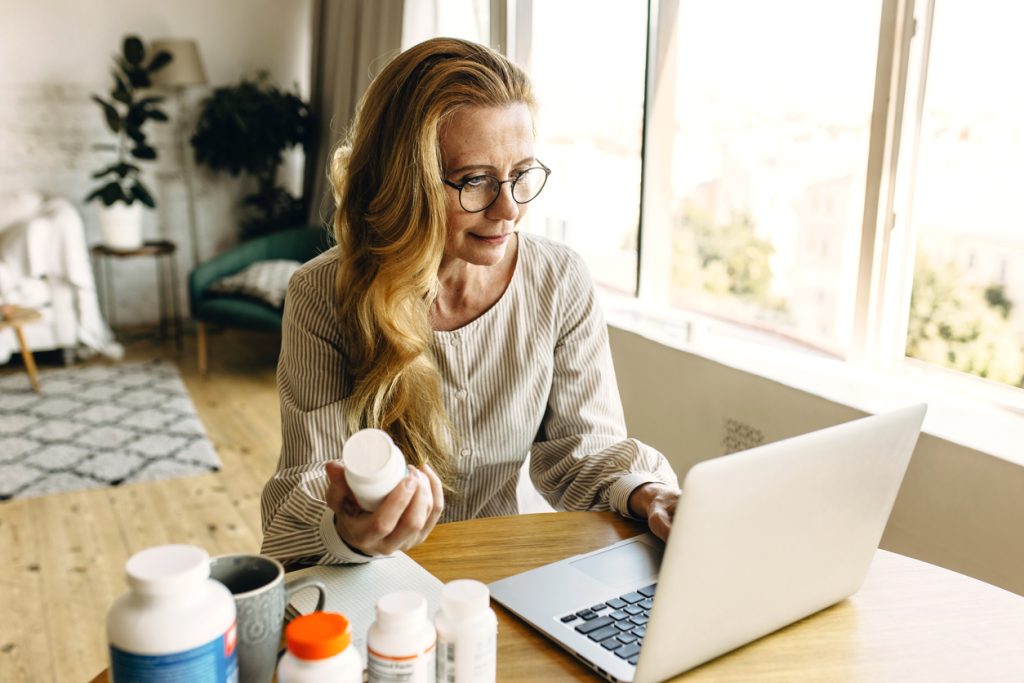 Kobieta w średnim wieku przy biurku czytająca napis na opakowaniu leku wspomagając się wiadomościami z internetu